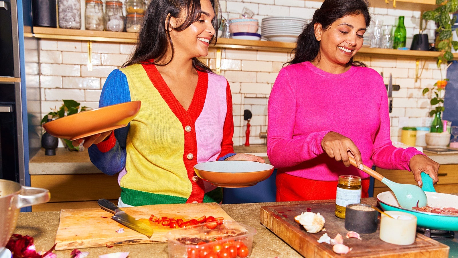 Two women smiling and cooking together in a kitchen, holding plates and preparing food on a bench.