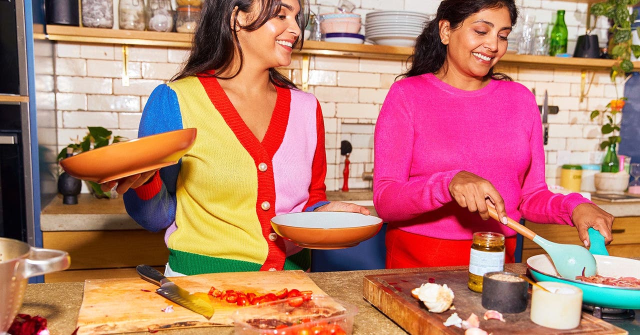 Two women smiling and cooking together in a kitchen, holding plates and preparing food on a bench.