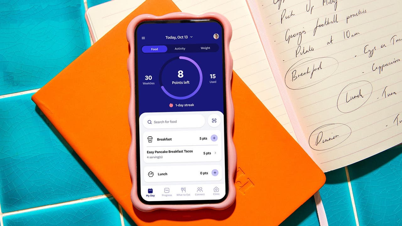 Close-up of a smartphone with the WeightWatchers app open, showing daily points tracking, placed on an orange notebook with handwritten notes in the background.