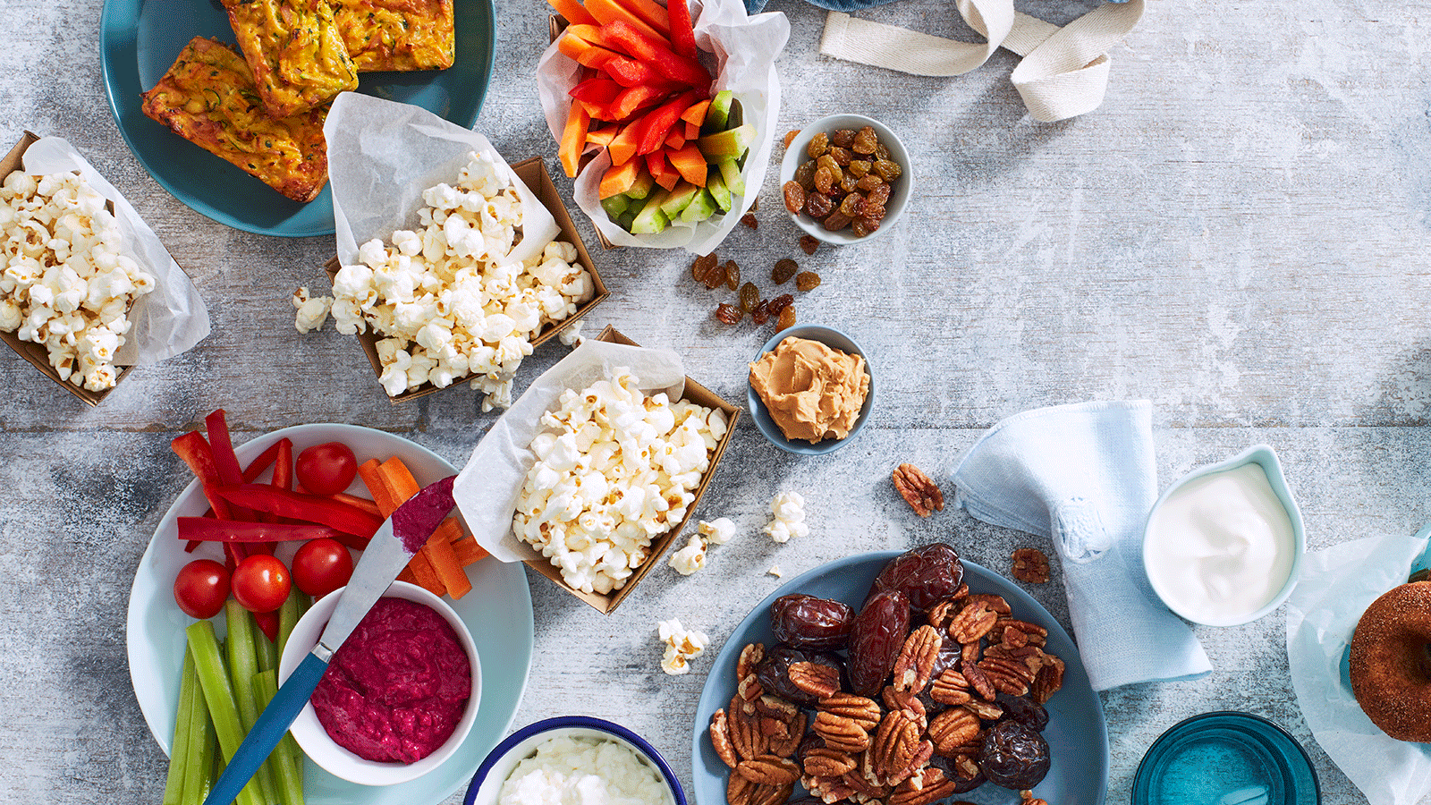 Healthy snack platter featuring popcorn, sliced vegetables with dip, nuts, dates, and mini zucchini slices.