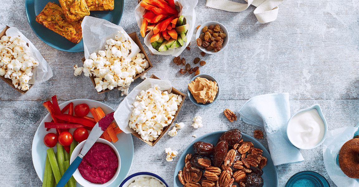 Healthy snack platter featuring popcorn, sliced vegetables with dip, nuts, dates, and mini zucchini slices.