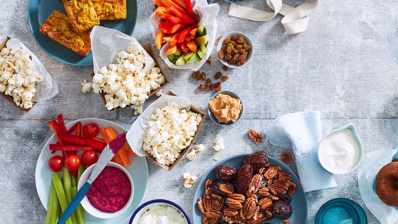 Healthy snack platter featuring popcorn, sliced vegetables with dip, nuts, dates, and mini zucchini slices.