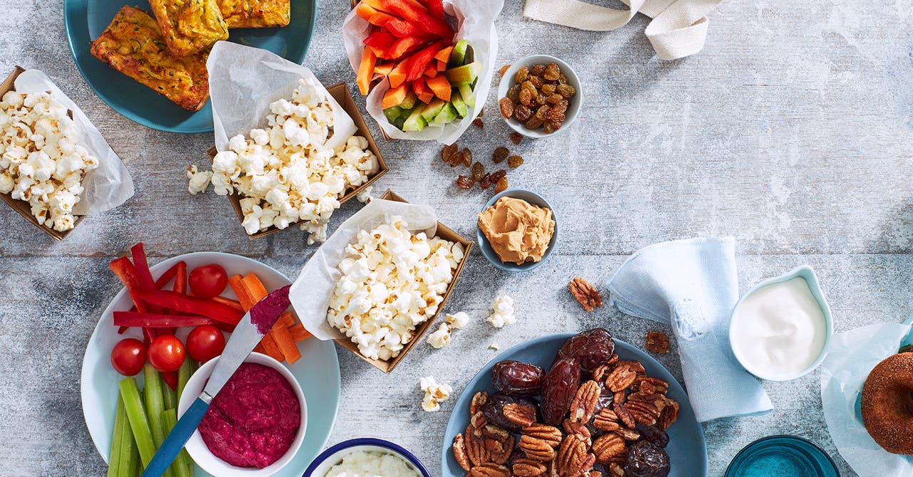 Healthy snack platter featuring popcorn, sliced vegetables with dip, nuts, dates, and mini zucchini slices.