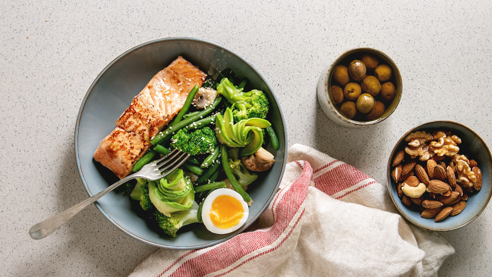 A healthy meal for managing PCOS with grilled salmon, green vegetables, and a soft-boiled egg. Side dishes include green olives and mixed nuts (almonds, walnuts, cashews).