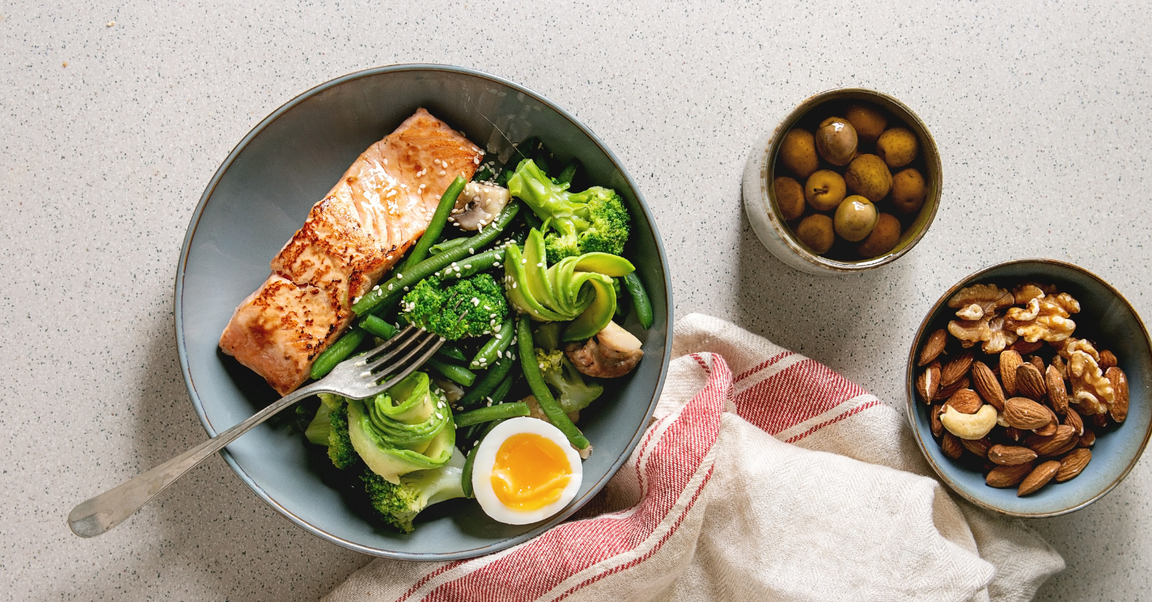 A healthy meal for managing PCOS with grilled salmon, green vegetables, and a soft-boiled egg. Side dishes include green olives and mixed nuts (almonds, walnuts, cashews).
