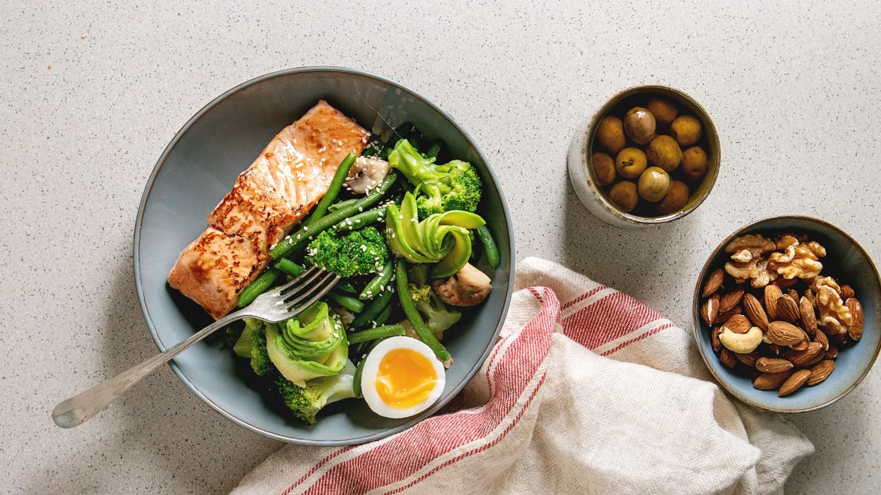 A healthy meal for managing PCOS with grilled salmon, green vegetables, and a soft-boiled egg. Side dishes include green olives and mixed nuts (almonds, walnuts, cashews).