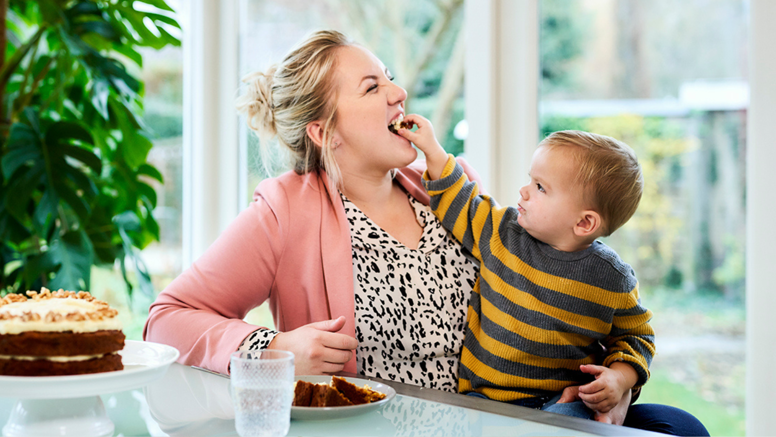 Mother laughing as her young child feeds her a bite of cake, with a slice of cake and a glass of water on the table.