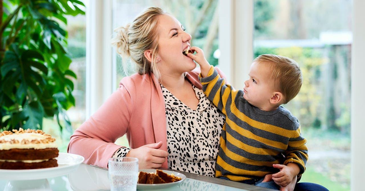 Mother laughing as her young child feeds her a bite of cake, with a slice of cake and a glass of water on the table.