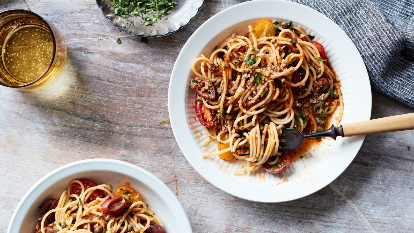 Plate of spaghetti alla puttanesca garnished with fresh herbs, accompanied by a glass of sparkling drink and a bowl of chopped herbs, set on a rustic wooden table.