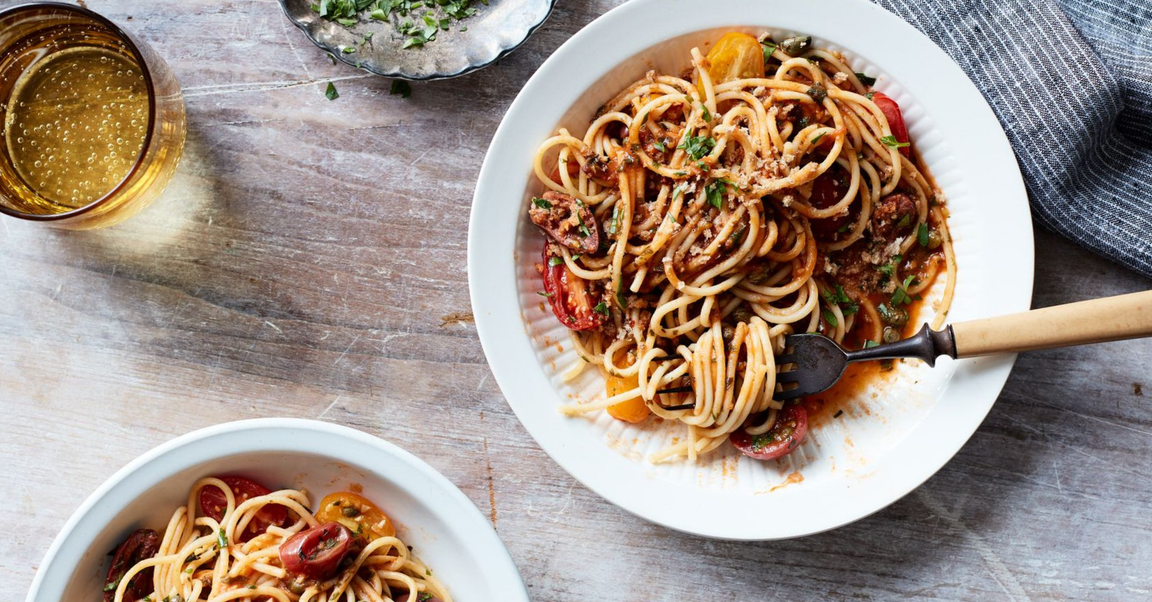 Plate of spaghetti alla puttanesca garnished with fresh herbs, accompanied by a glass of sparkling drink and a bowl of chopped herbs, set on a rustic wooden table.