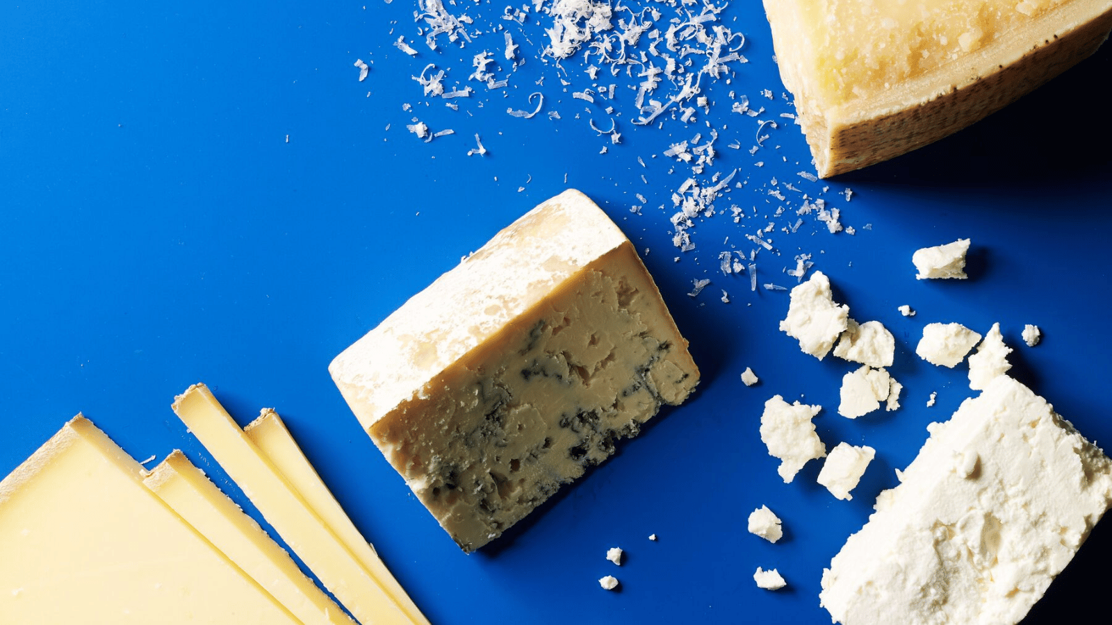 Various types of cheese on a bright blue background, featuring both hard and crumbly varieties.