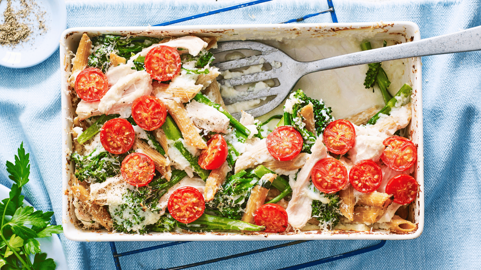 A baked chicken and pasta dish topped with fresh cherry tomatoes and leafy greens, served in a white baking dish with a blue background.