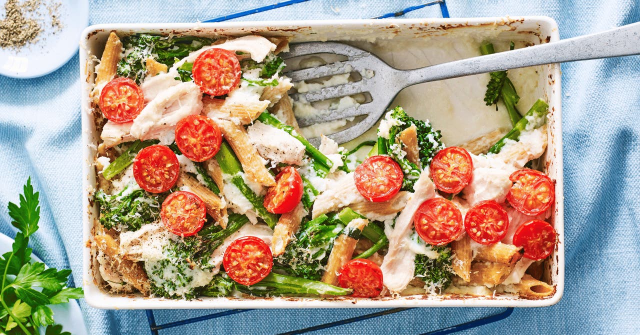 A baked chicken and pasta dish topped with fresh cherry tomatoes and leafy greens, served in a white baking dish with a blue background.