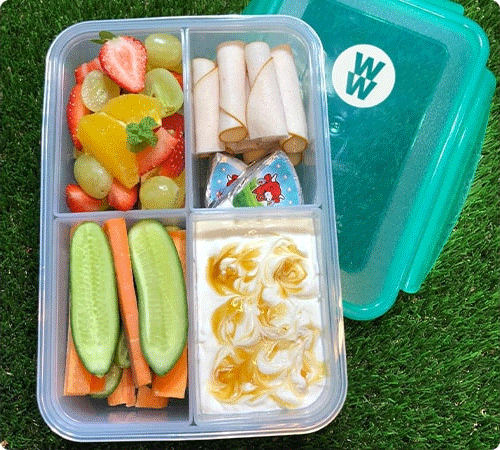 A bento box with fruit, turkey slices, cucumber and carrot sticks, and yogurt with honey. The box has a teal WeightWatchers lid.