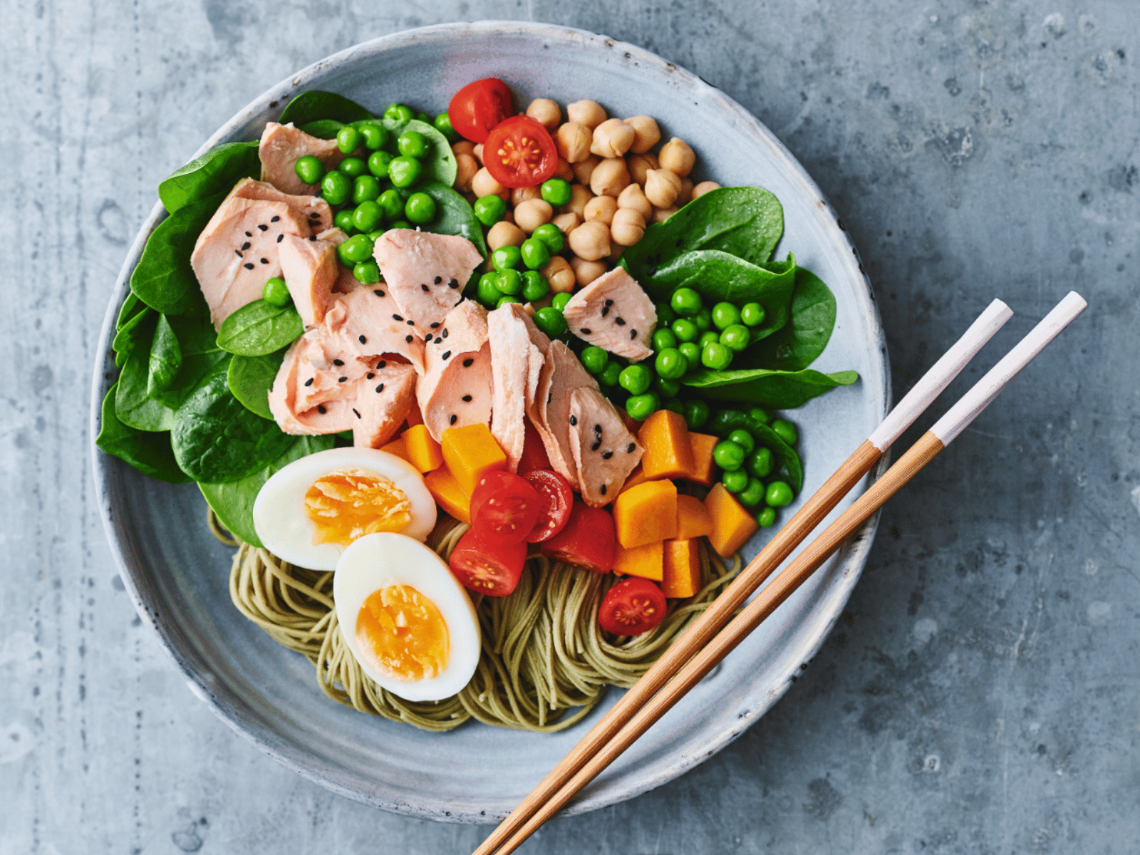 A colourful salad bowl with spinach, peas, chickpeas, cherry tomatoes, boiled eggs, sliced salmon with black sesame seeds, and soba noodles. The dish is served with chopsticks.