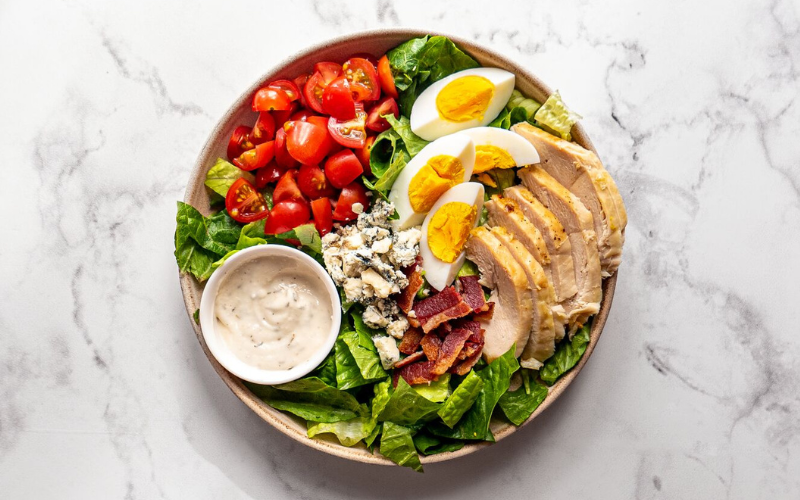 A healthy salad bowl featuring fresh romaine lettuce, cherry tomatoes, blue cheese crumbles, crispy bacon, hard-boiled eggs, and sliced grilled chicken breast, accompanied by a side of creamy dressing.