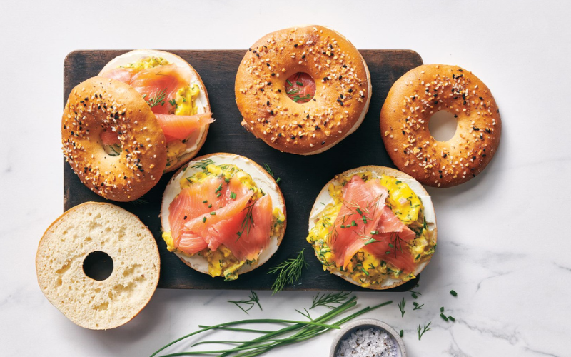 Everything bagels topped with scrambled eggs and smoked salmon, garnished with fresh dill, placed on a wooden board on a marble surface.