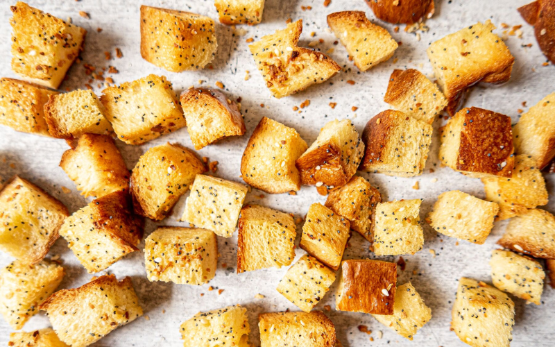 Close-up of crispy, golden-brown everything bagel croutons scattered on a white surface.