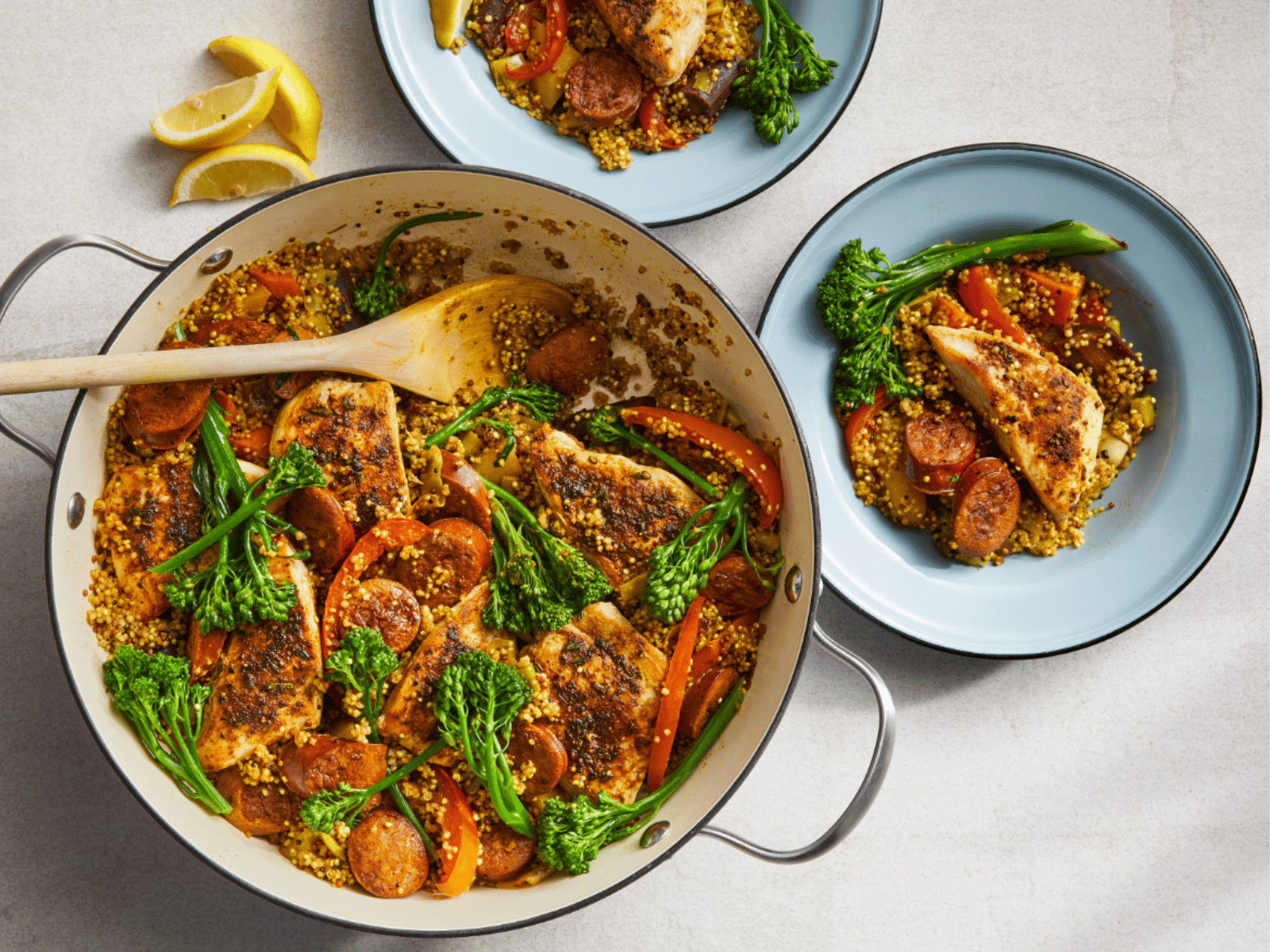 A vibrant dish of seasoned chicken, chorizo, and vegetables including broccolini and bell peppers, served on a bed of grains in a large pan with lemon wedges on the side.