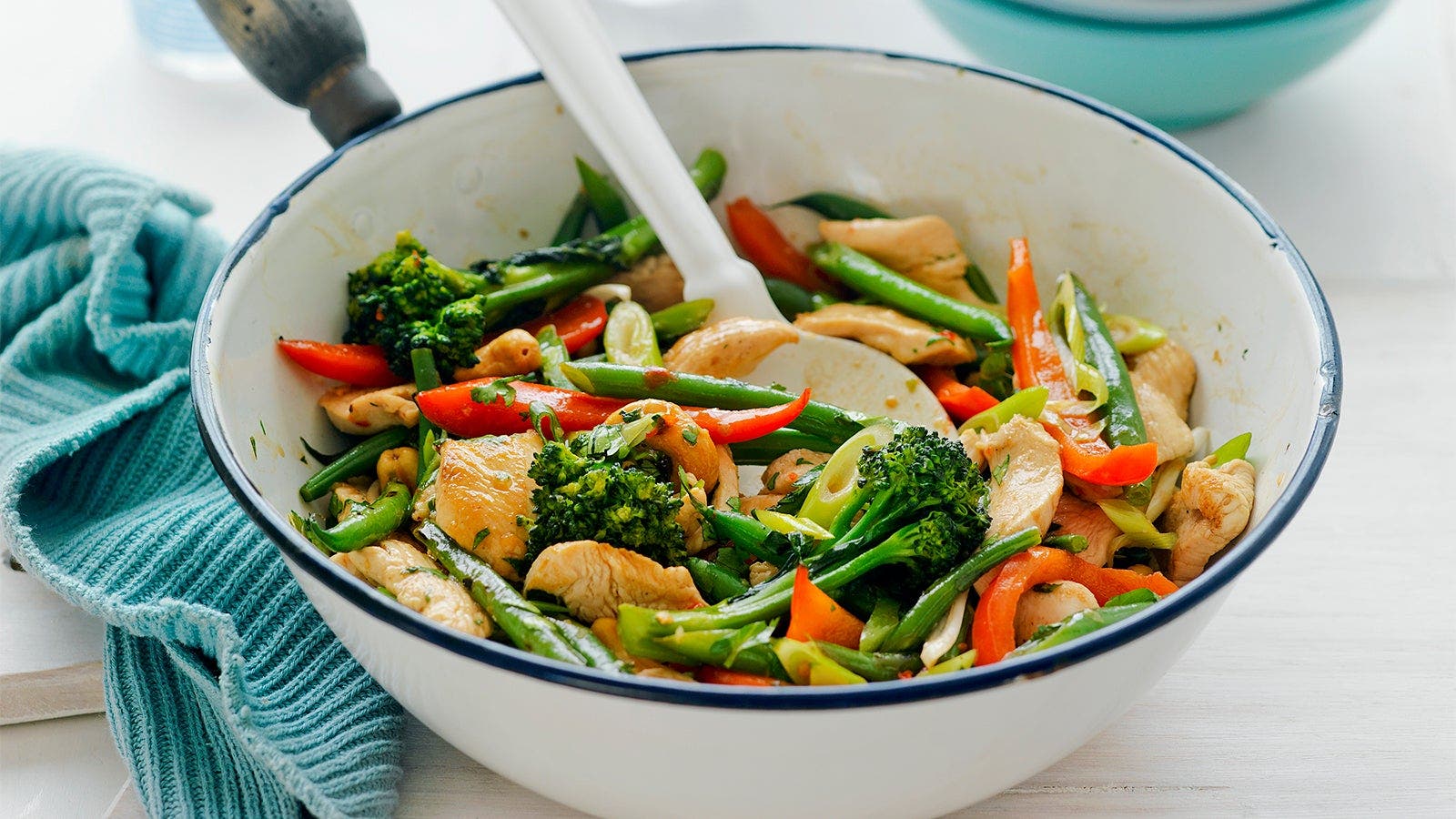 A colourful chicken stir-fry with a variety of fresh vegetables including broccoli, red capsicum, and green beans in a white pot. A white spoon is used for serving.