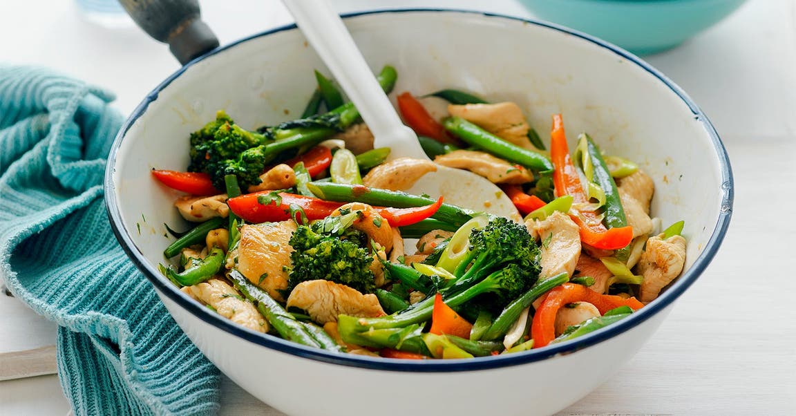 A colourful chicken stir-fry with a variety of fresh vegetables including broccoli, red capsicum, and green beans in a white pot. A white spoon is used for serving.
