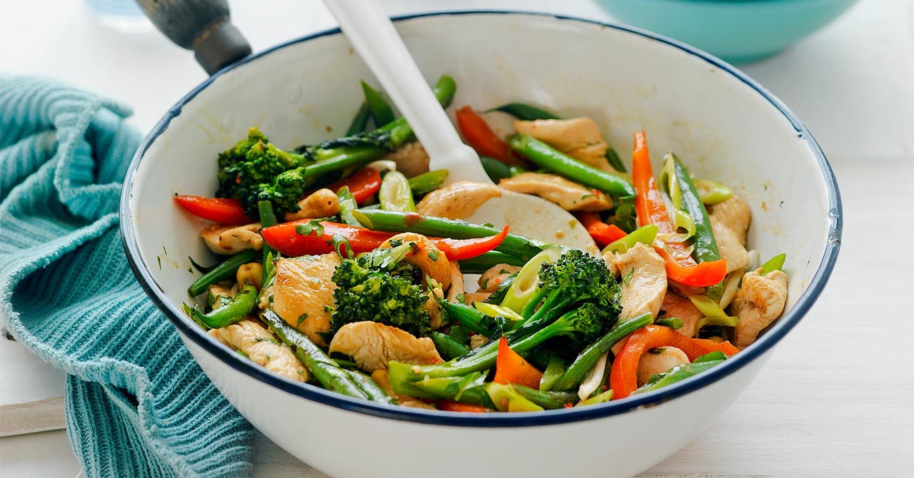A colourful chicken stir-fry with a variety of fresh vegetables including broccoli, red capsicum, and green beans in a white pot. A white spoon is used for serving.