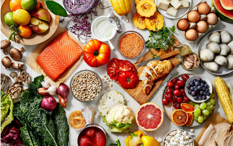 A colourful assortment of fresh fruits, vegetables, and proteins including salmon, chicken breast, eggs, tofu, legumes, nuts, and various produce items such as tomatoes, grapes, oranges, and leafy greens.