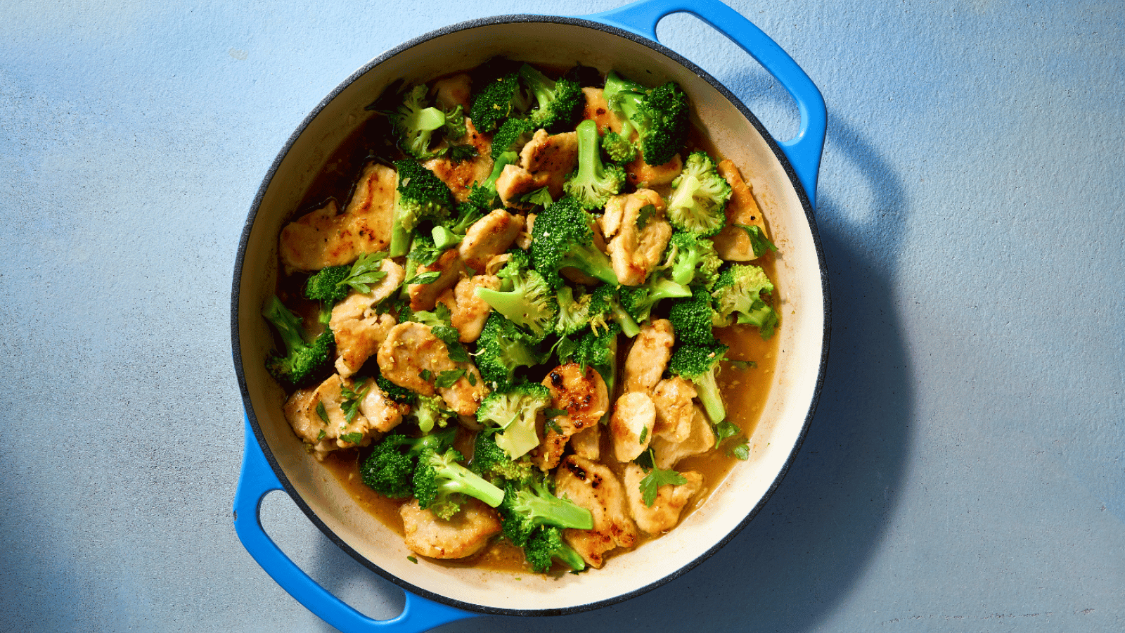 A pan of chicken and broccoli stir-fry in a light sauce, garnished with parsley, served in a blue-handled pot.