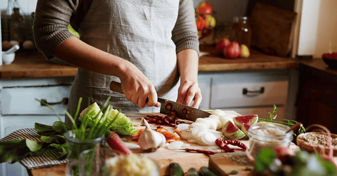 Person chopping fresh vegetables including onions, lettuce, and radishes on a wooden kitchen counter, surrounded by various ingredients.