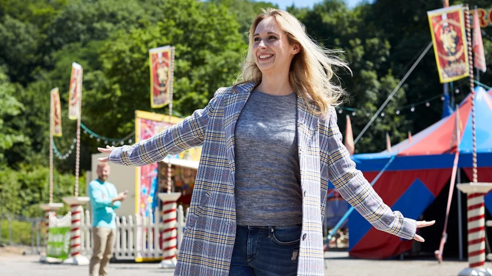 A smiling woman in a plaid coat with arms outstretched at an outdoor carnival.