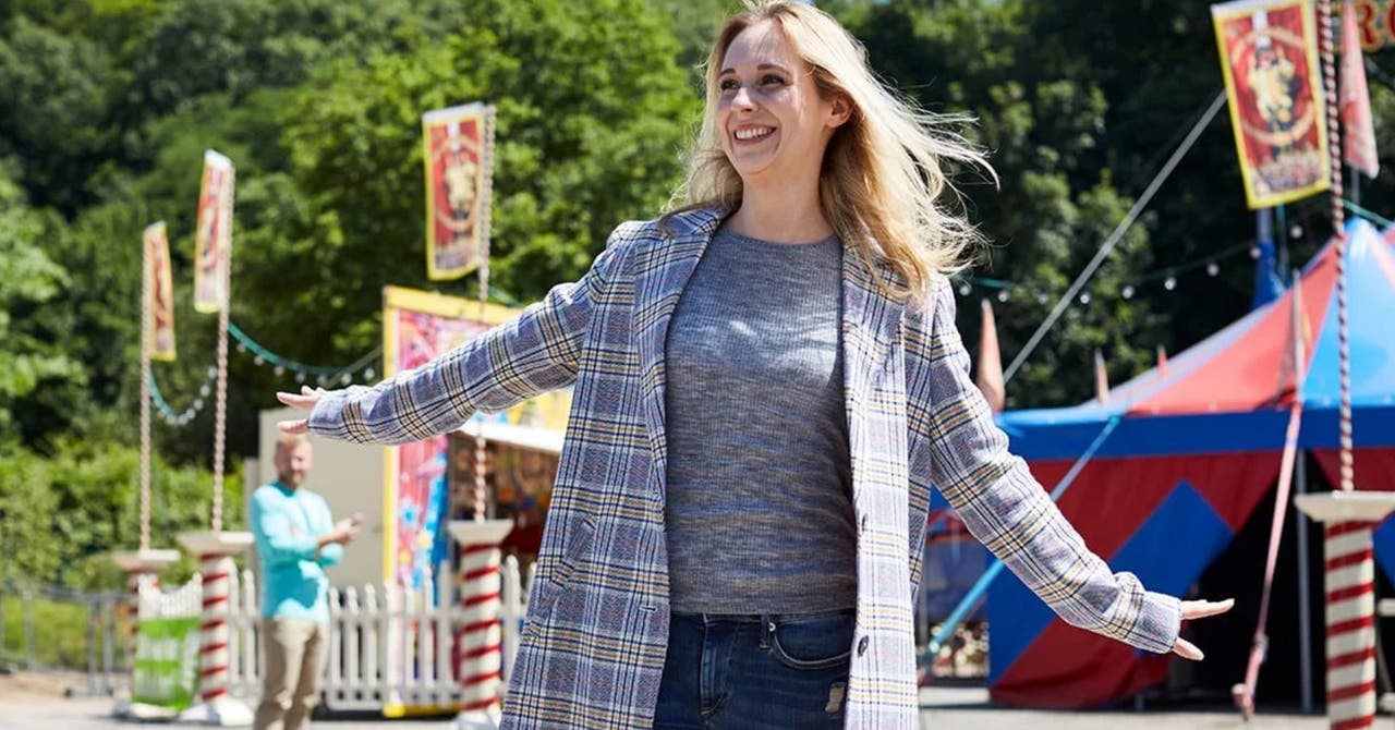 A smiling woman in a plaid coat with arms outstretched at an outdoor carnival.
