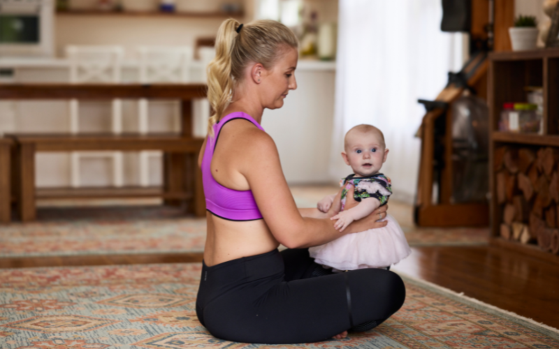 A woman in workout attire sitting on the floor and holding a baby dressed in a cute outfit.