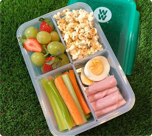 A bento box with grapes, strawberries, popcorn, veggie sticks, a boiled egg, and ham slices. The box has a teal WeightWatchers lid.