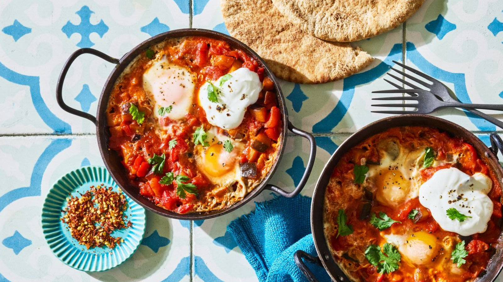 Two pans of shakshuka, a dish with poached eggs in a spiced tomato and vegetable sauce, served with pita bread and a small dish of red pepper flakes.