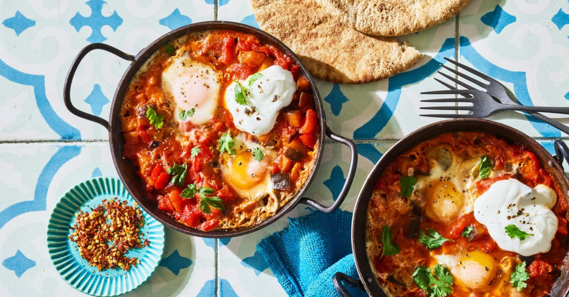 Two pans of shakshuka, a dish with poached eggs in a spiced tomato and vegetable sauce, served with pita bread and a small dish of red pepper flakes.