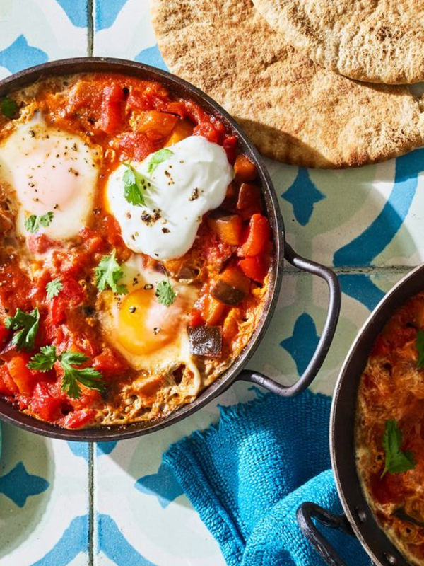 Two pans of shakshuka, a dish with poached eggs in a spiced tomato and vegetable sauce, served with pita bread and a small dish of red pepper flakes.