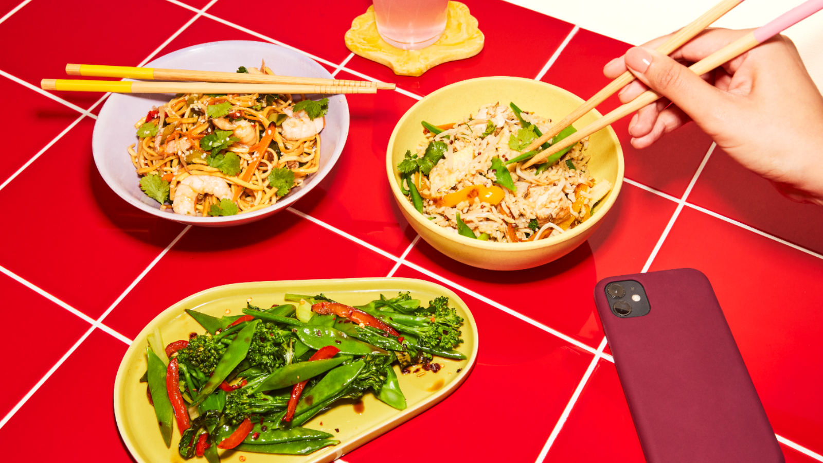 An assortment of Asian-inspired dishes including stir-fried noodles with prawns, fried rice with vegetables, and a plate of mixed greens, placed on a red tiled table.