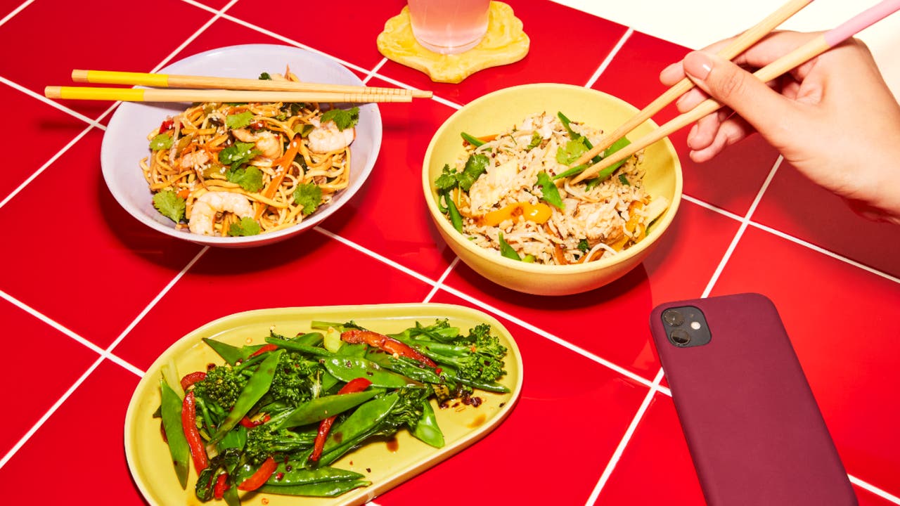 An assortment of Asian-inspired dishes including stir-fried noodles with prawns, fried rice with vegetables, and a plate of mixed greens, placed on a red tiled table.