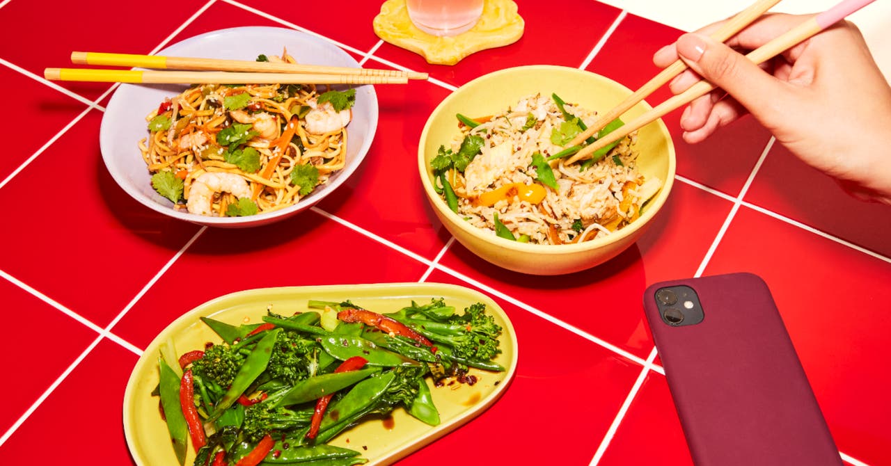 An assortment of Asian-inspired dishes including stir-fried noodles with prawns, fried rice with vegetables, and a plate of mixed greens, placed on a red tiled table.