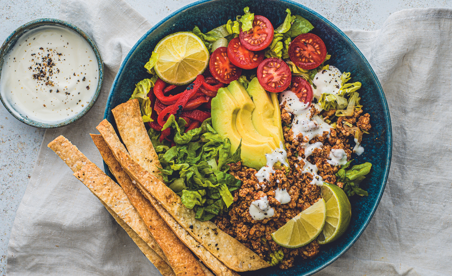 Healthy taco salad bowl featuring crisp lettuce, sliced avocado, cherry tomatoes, lime wedges, seasoned ground meat, tortilla strips, and a side of creamy dressing.