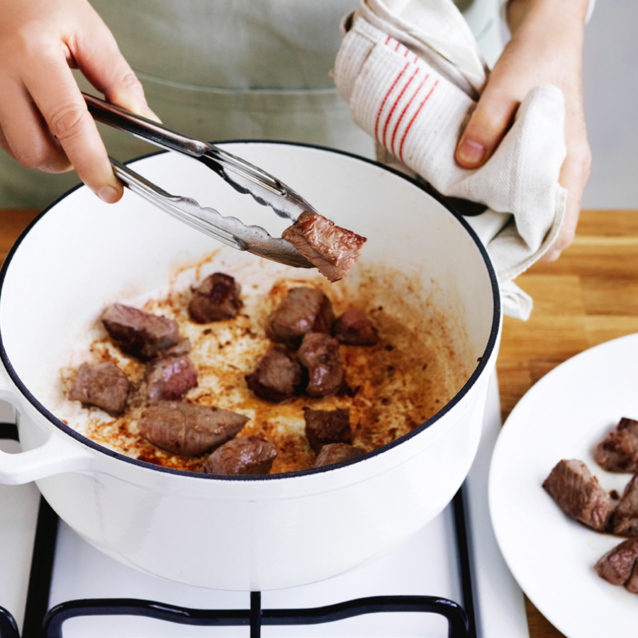 A person browning meat in a white pot on a stovetop, preparing the base for a stew.