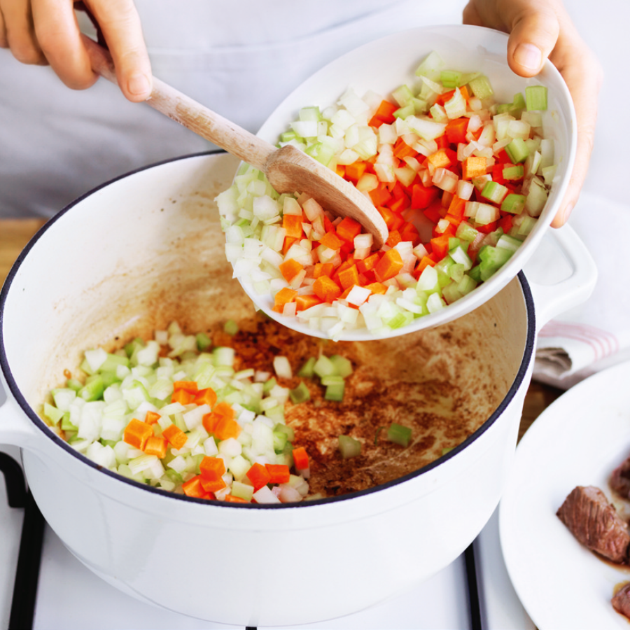 A person adding a mix of chopped onions, carrots, and celery into a white pot on a stovetop, preparing the base for a stew.