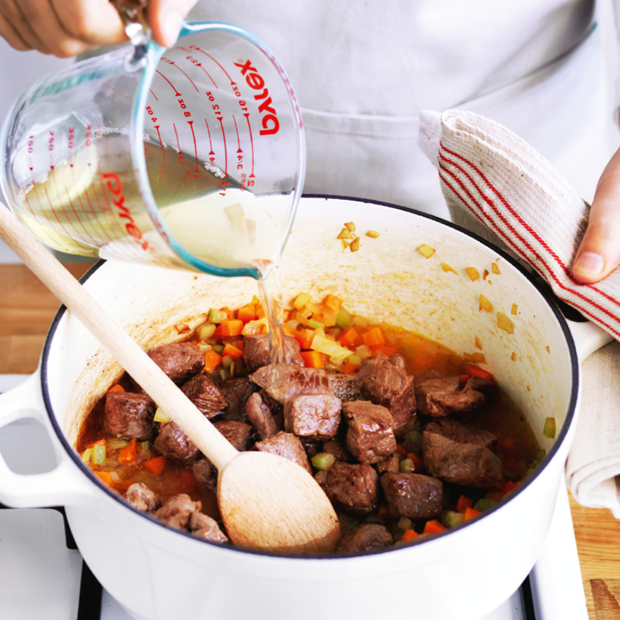 A person adding stock into a white pot on a stovetop, preparing the base for a stew.