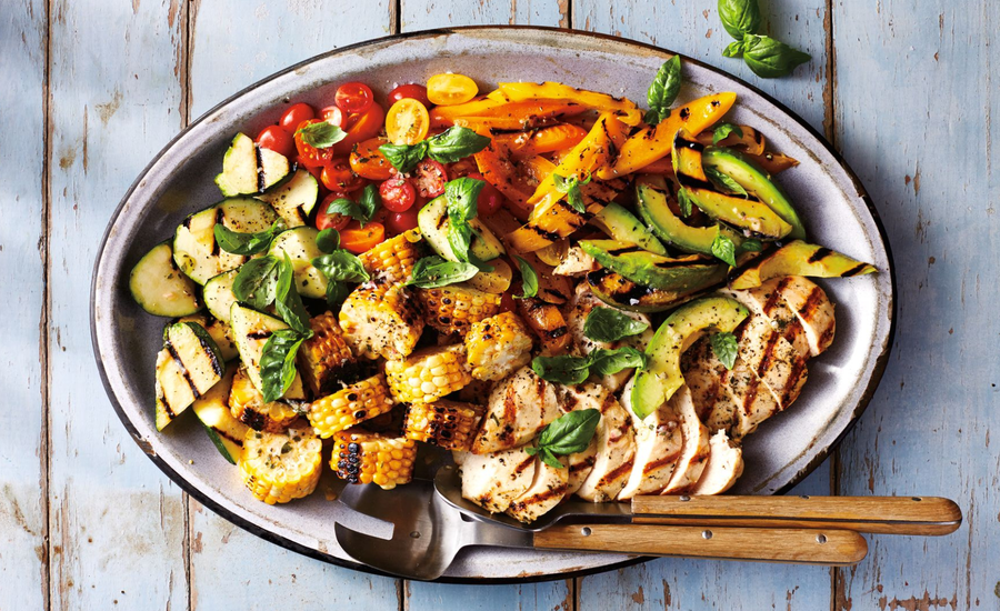 Grilled chicken and vegetable platter featuring sliced grilled chicken breast, charred corn, zucchini, cherry tomatoes, capsicum and avocado, garnished with fresh basil.