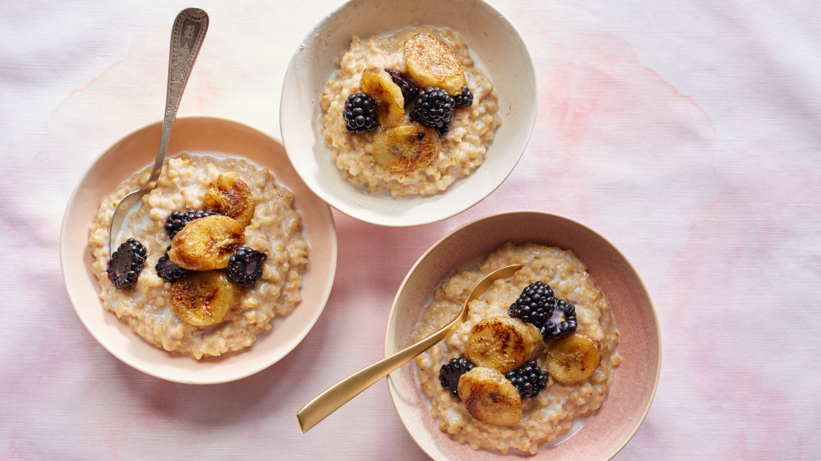 Three bowls of oats topped with caramelised banana slices and blackberries on a light pink background.