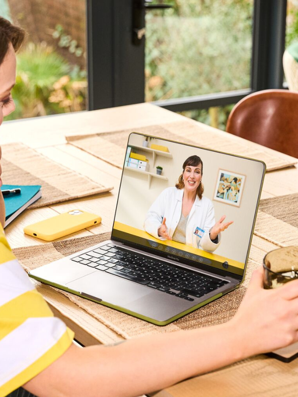 A woman having a virtual consultation on a laptop with a female doctor, while sitting at a table with a drink in her hand.