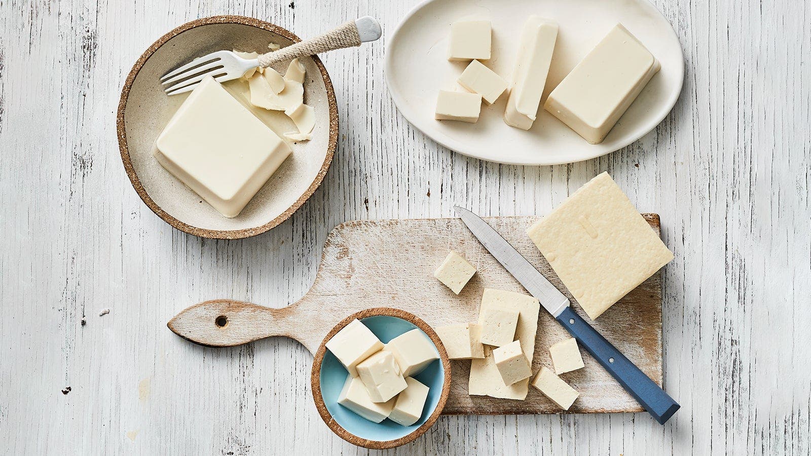 Assorted blocks and cubes of tofu displayed on different plates and a wooden cutting board, with a knife and a fork.