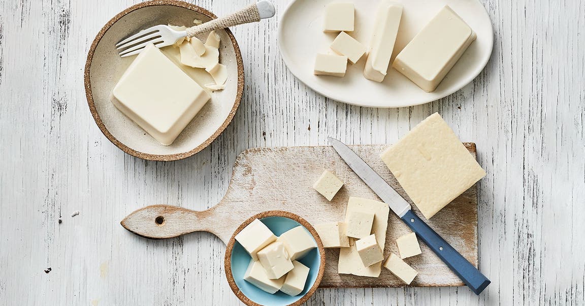 Assorted blocks and cubes of tofu displayed on different plates and a wooden cutting board, with a knife and a fork.