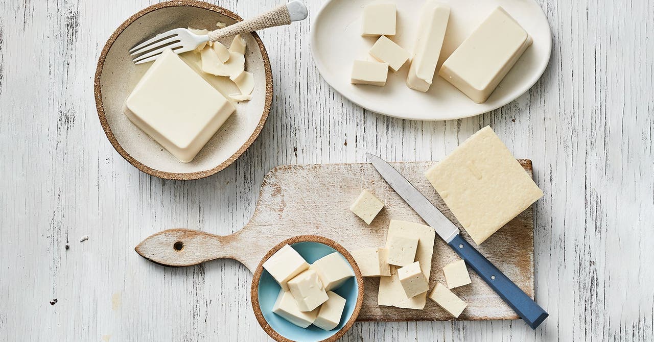 Assorted blocks and cubes of tofu displayed on different plates and a wooden cutting board, with a knife and a fork.