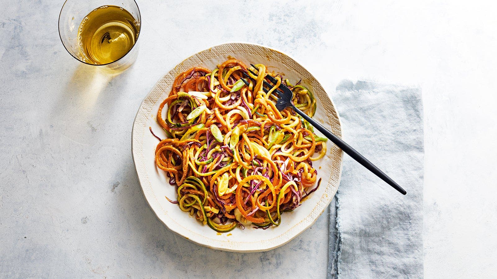 A plate of spiralized vegetables, including carrots and zucchini, garnished with sliced green onions, next to a glass of light-coloured drink.